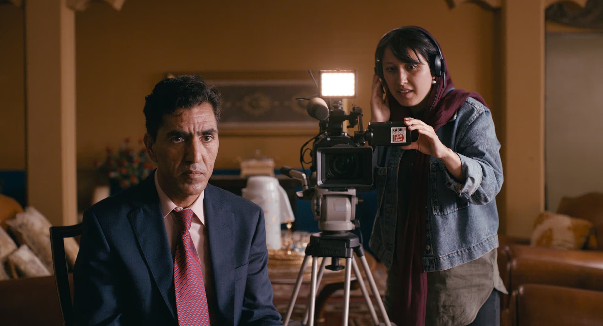 A woman with headphones films a man in a suit with a professional camera indoors.