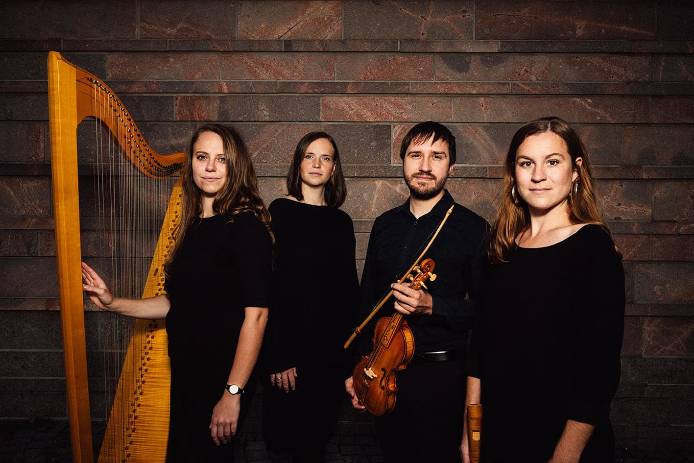 Three musicians and a harpist pose in black clothes against a brown stone wall.