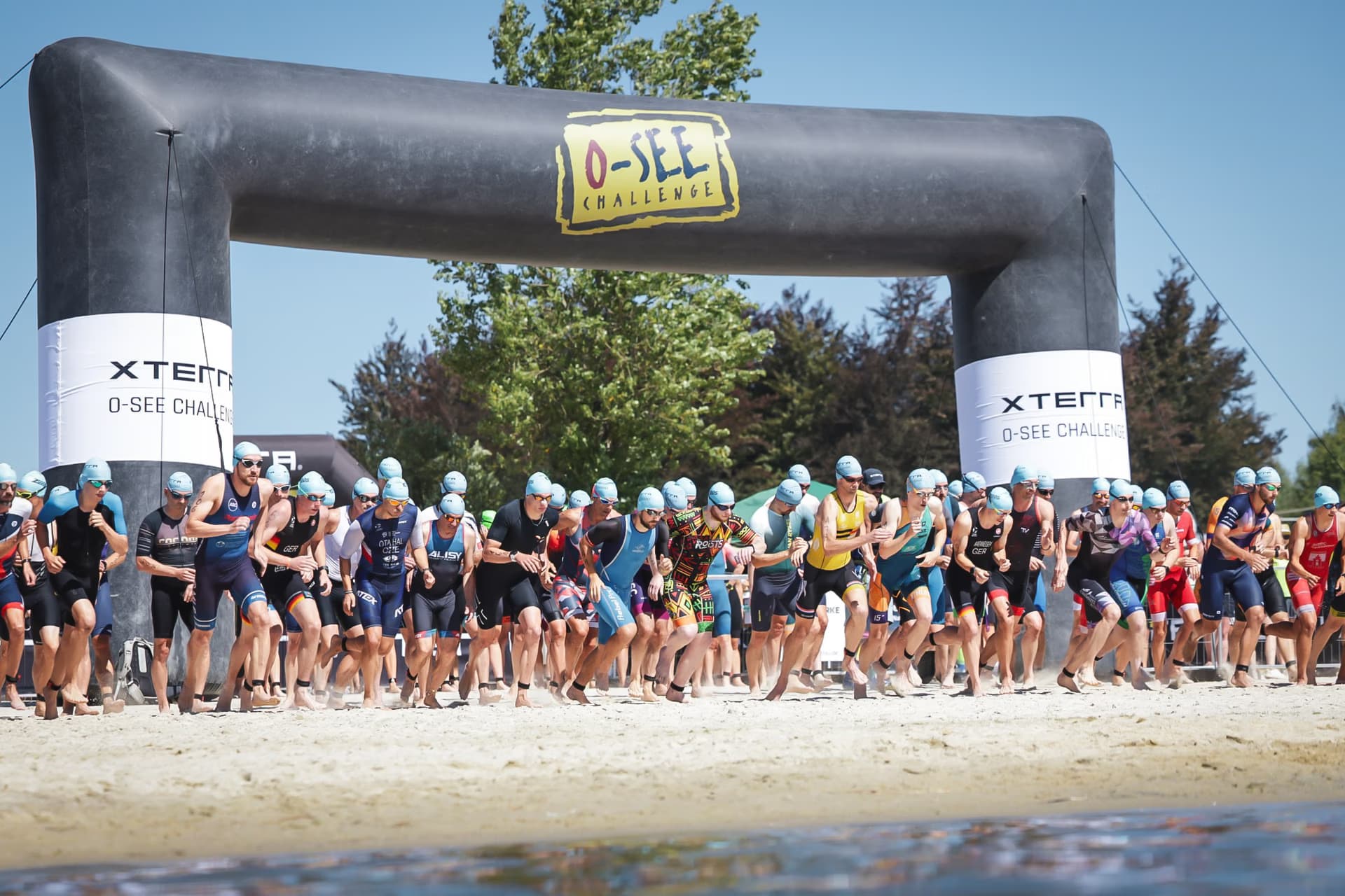 Athletes in swim gear run from a sandy beach under an O-SEE Challenge inflatable start arch.