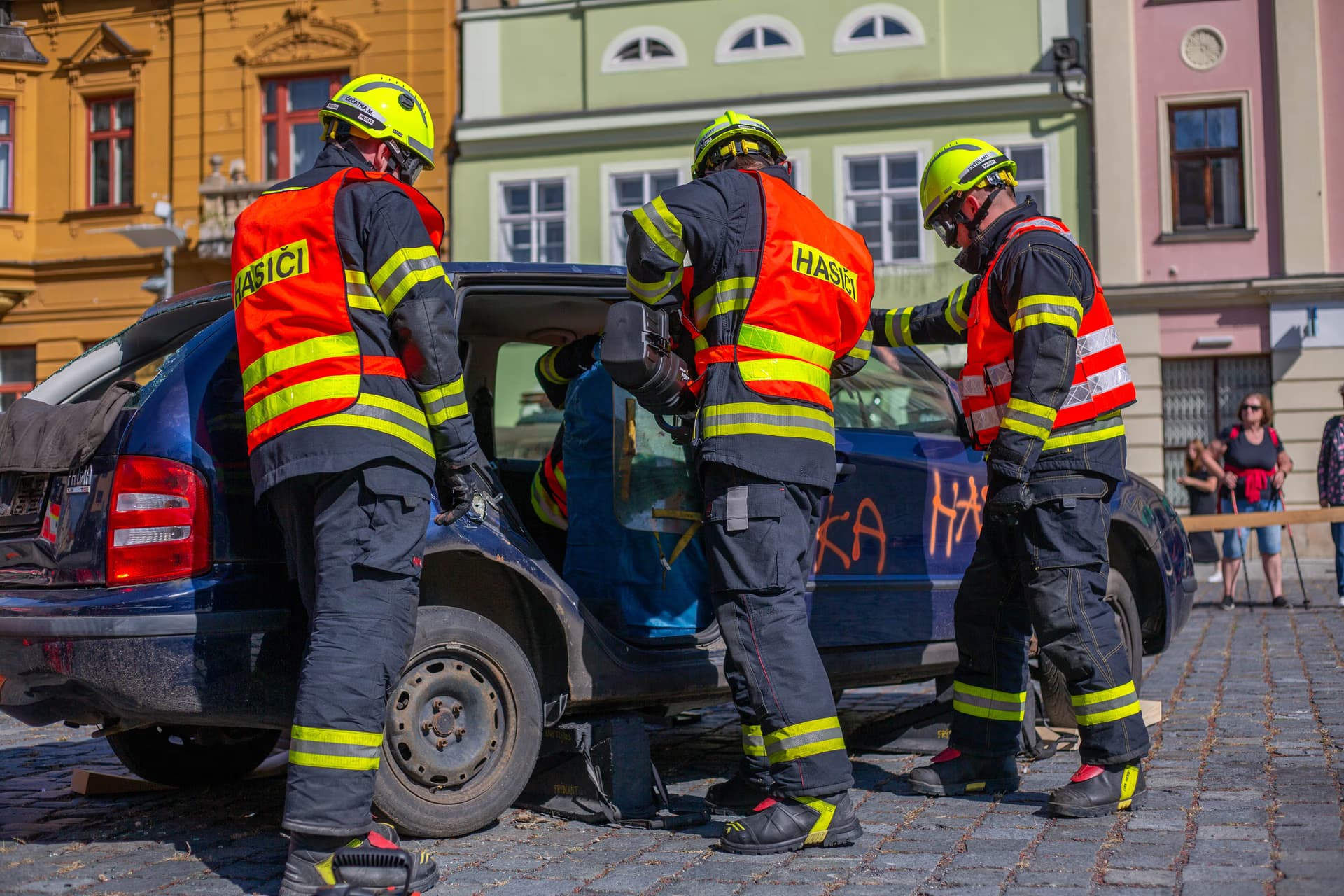 Tři hasiči v uniformách pracují na autě během ukázky na městském náměstí.