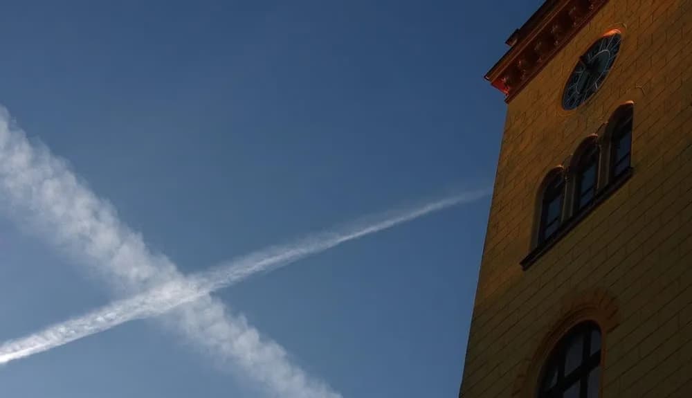 A portion of a yellow clock tower stands against a deep blue sky with crossing airplane contrails.