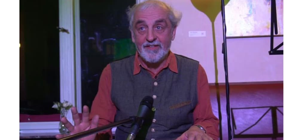 An older man with a grey beard speaks into a microphone in an indoor setting.