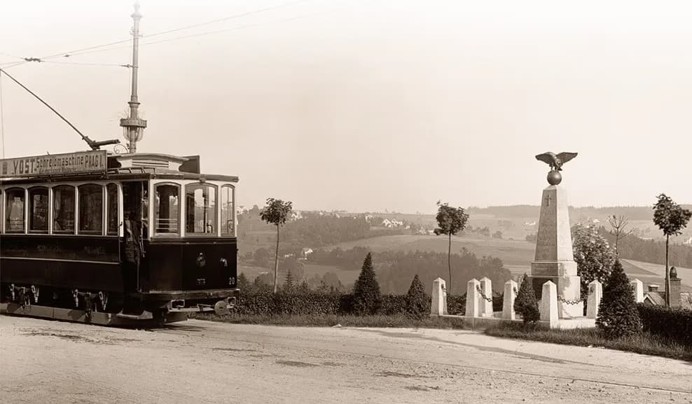 A vintage tram travels past a stone monument topped with an eagle statue in a hilly landscape.