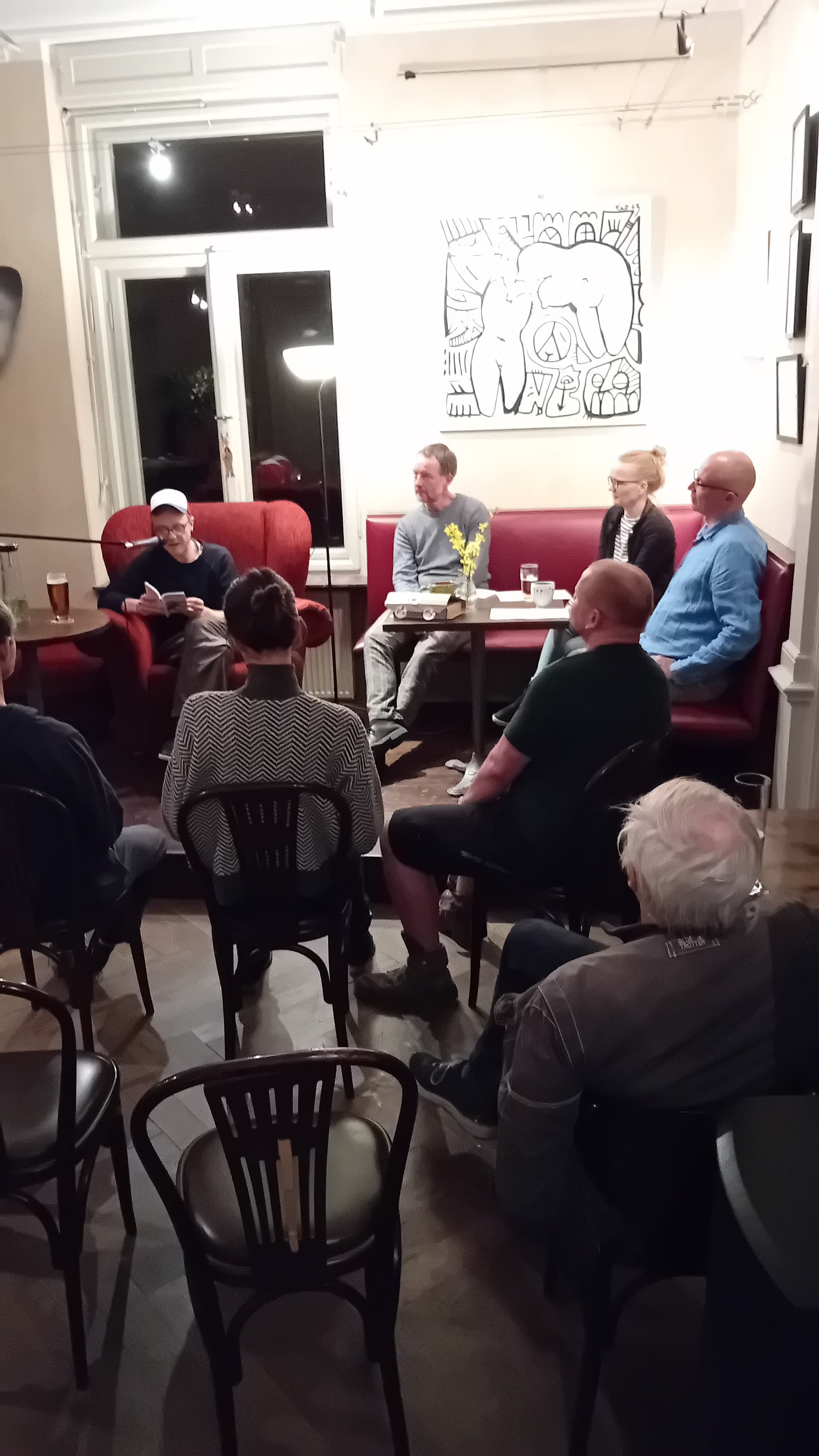 A group of people sits in a small cafe listening to a person reading aloud.