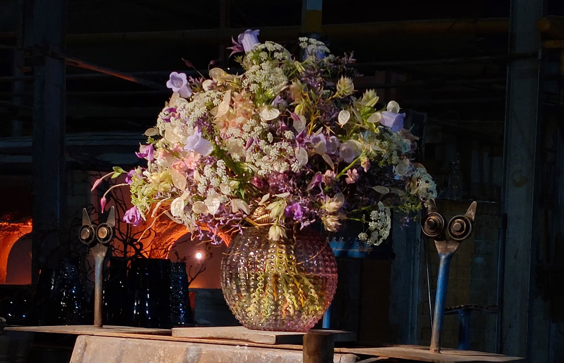 A lush, colorful bouquet of flowers sits in a textured glass vase inside a workshop.