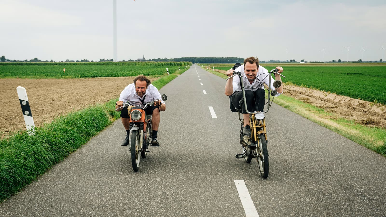 Two men in white shirts ride vintage mopeds along a quiet rural road between fields.