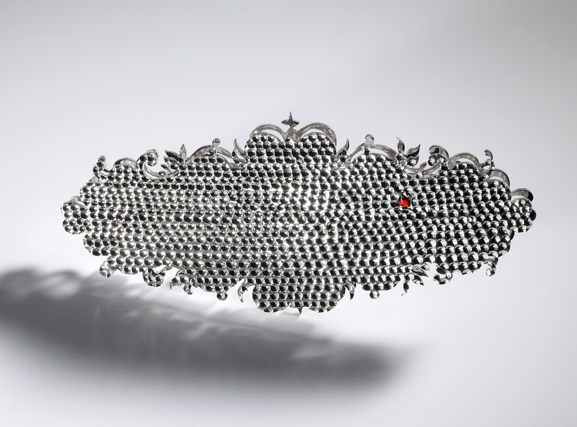 An ornate, elongated metallic decorative object covered in small studs floats against a light grey background.
