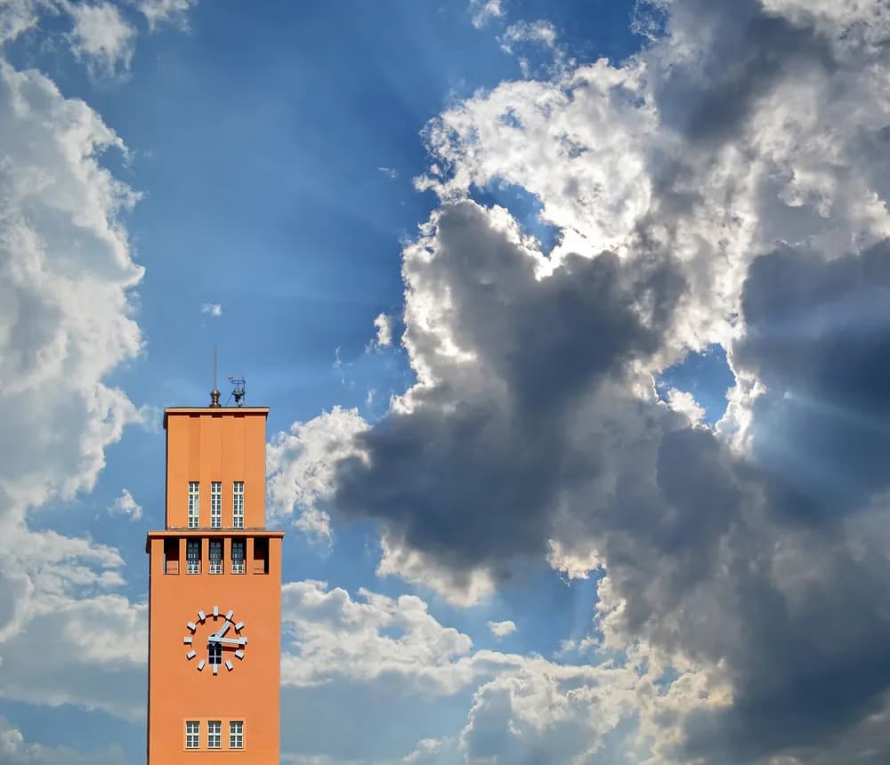 Orange clock tower stands against a bright blue sky with dramatic, fluffy white and dark clouds.