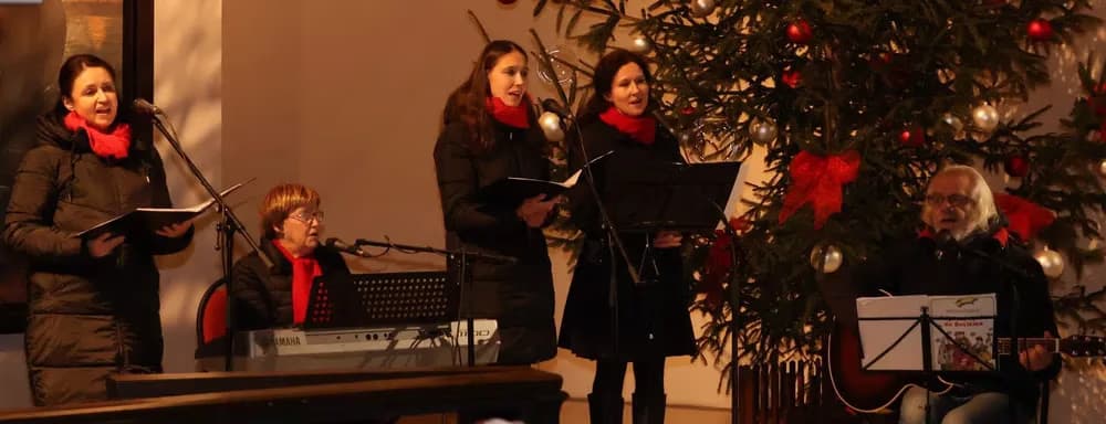 Trzy kobiety śpiewają, a mężczyzna gra na gitarze obok udekorowanej choinki wewnątrz pomieszczenia.
