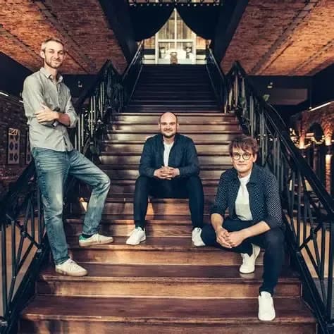 Three men posing on a grand wooden staircase inside an industrial-style building.