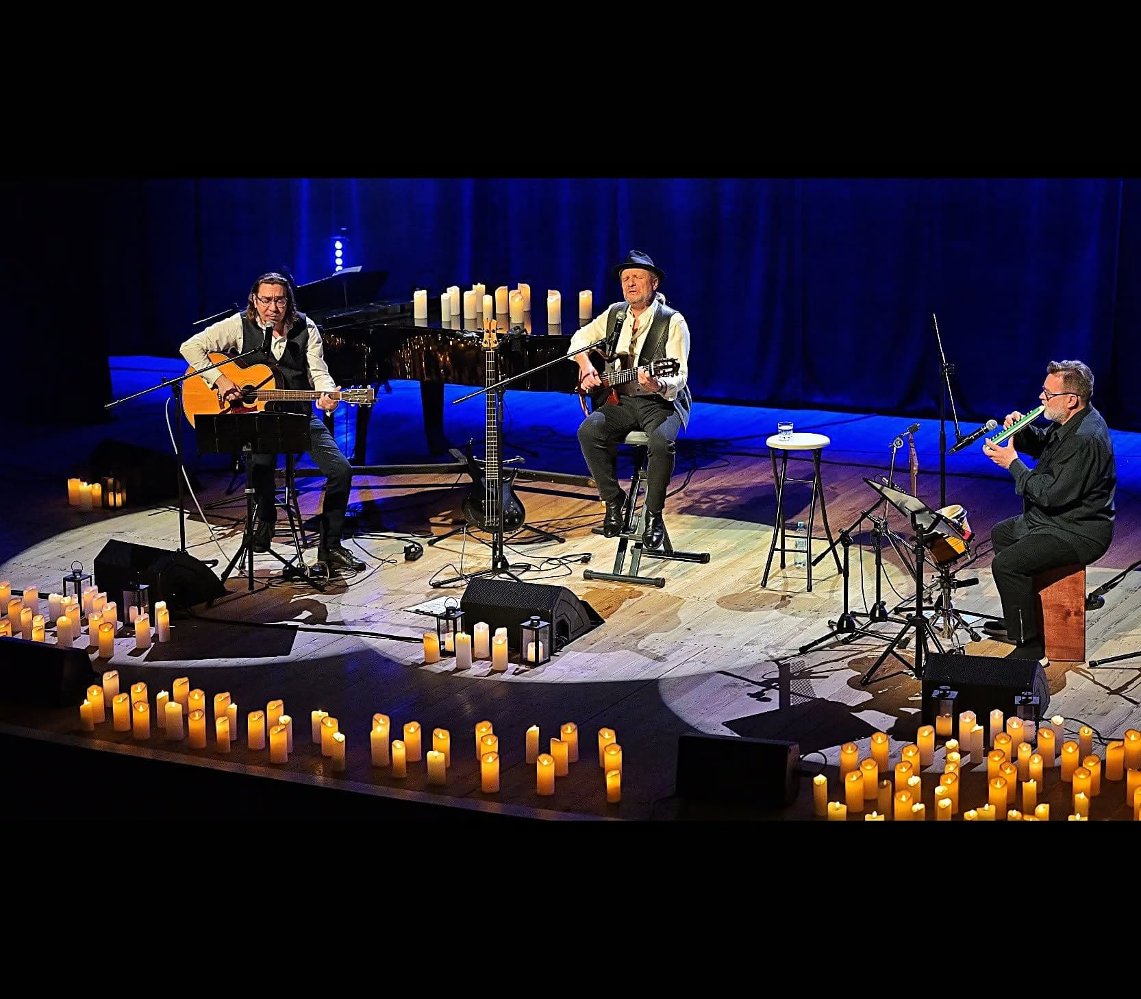 Three musicians perform on a stage surrounded by numerous lit candles in a dark room.
