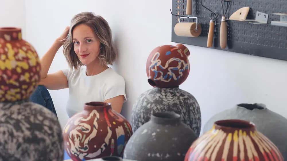 A smiling woman sits near several colorful handmade ceramic vases in a pottery studio.