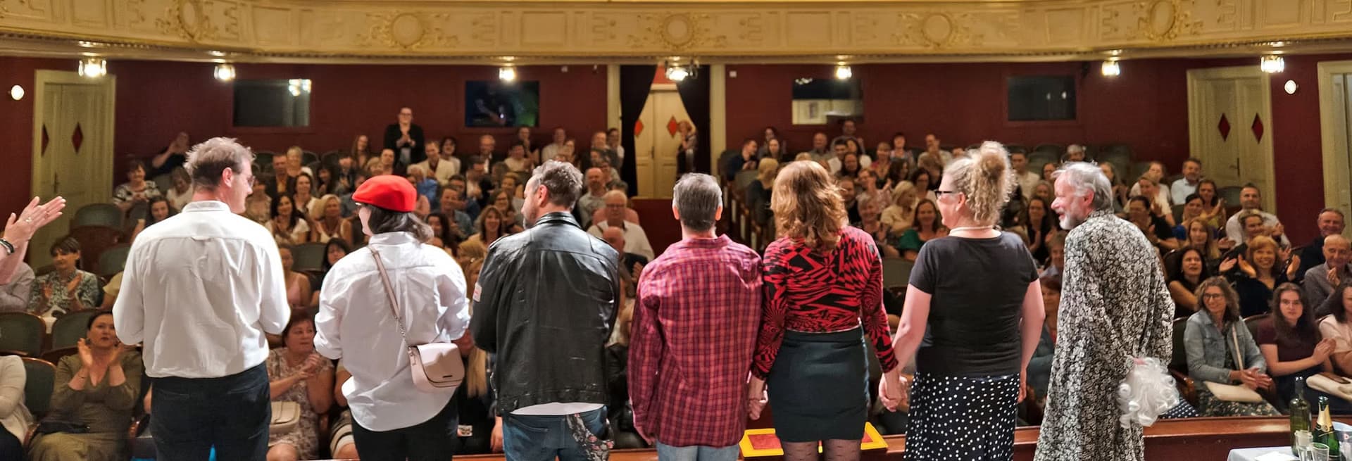 Mehrere Personen stehen auf einer Theaterbühne und blicken ins Publikum im vollen Saal.