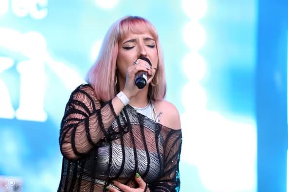 A pink-haired singer performs passionately into a microphone on an outdoor stage.