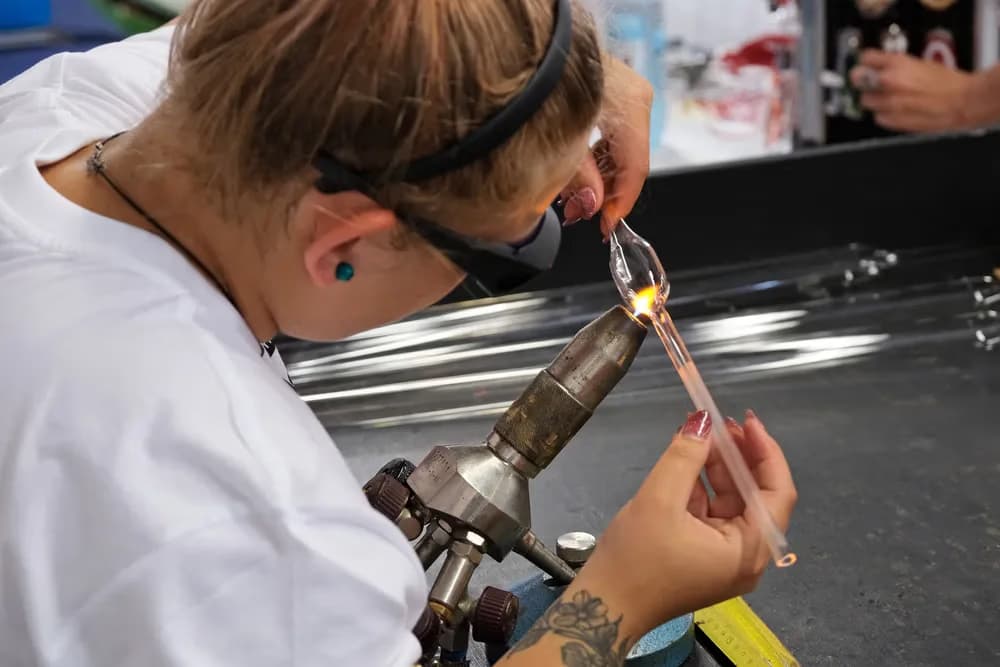 A glassblower wears protective glasses while shaping molten glass with a torch flame.