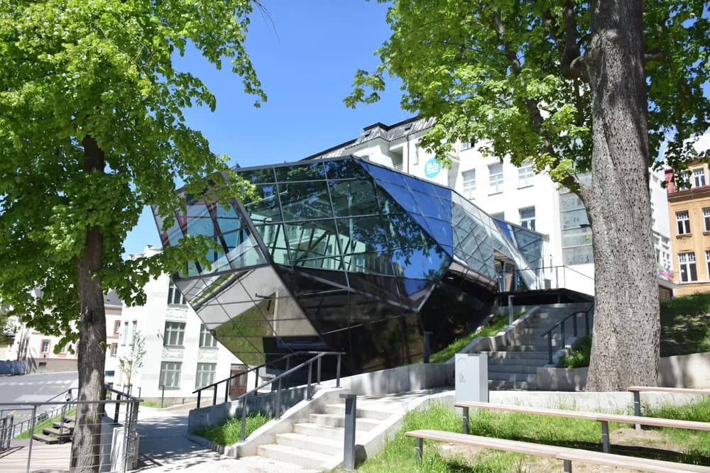 Modern angular glass architecture stands beside a white building surrounded by green trees on a sunny day.