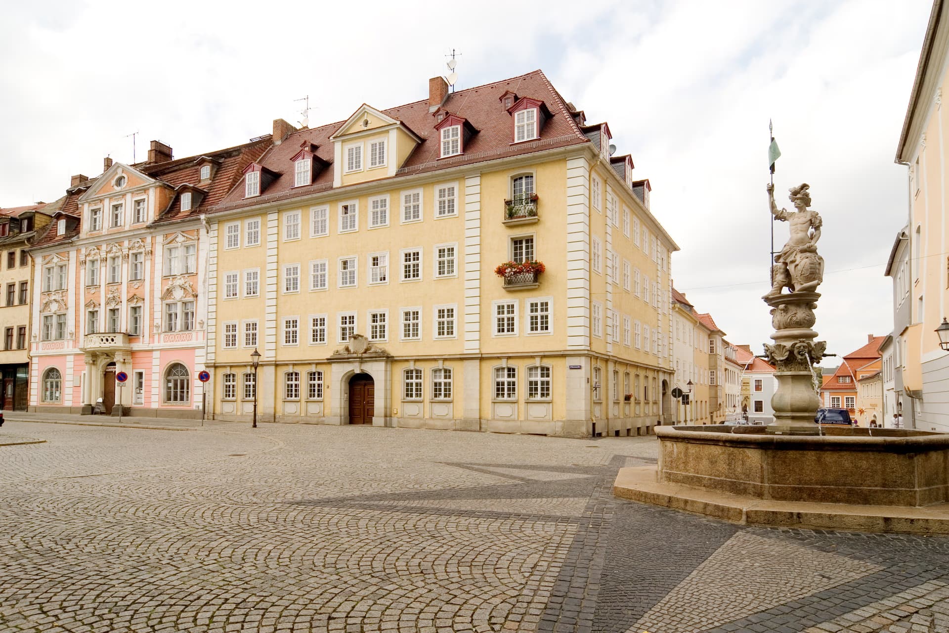 A cobblestone town square with historic, light-colored buildings and a stone fountain.