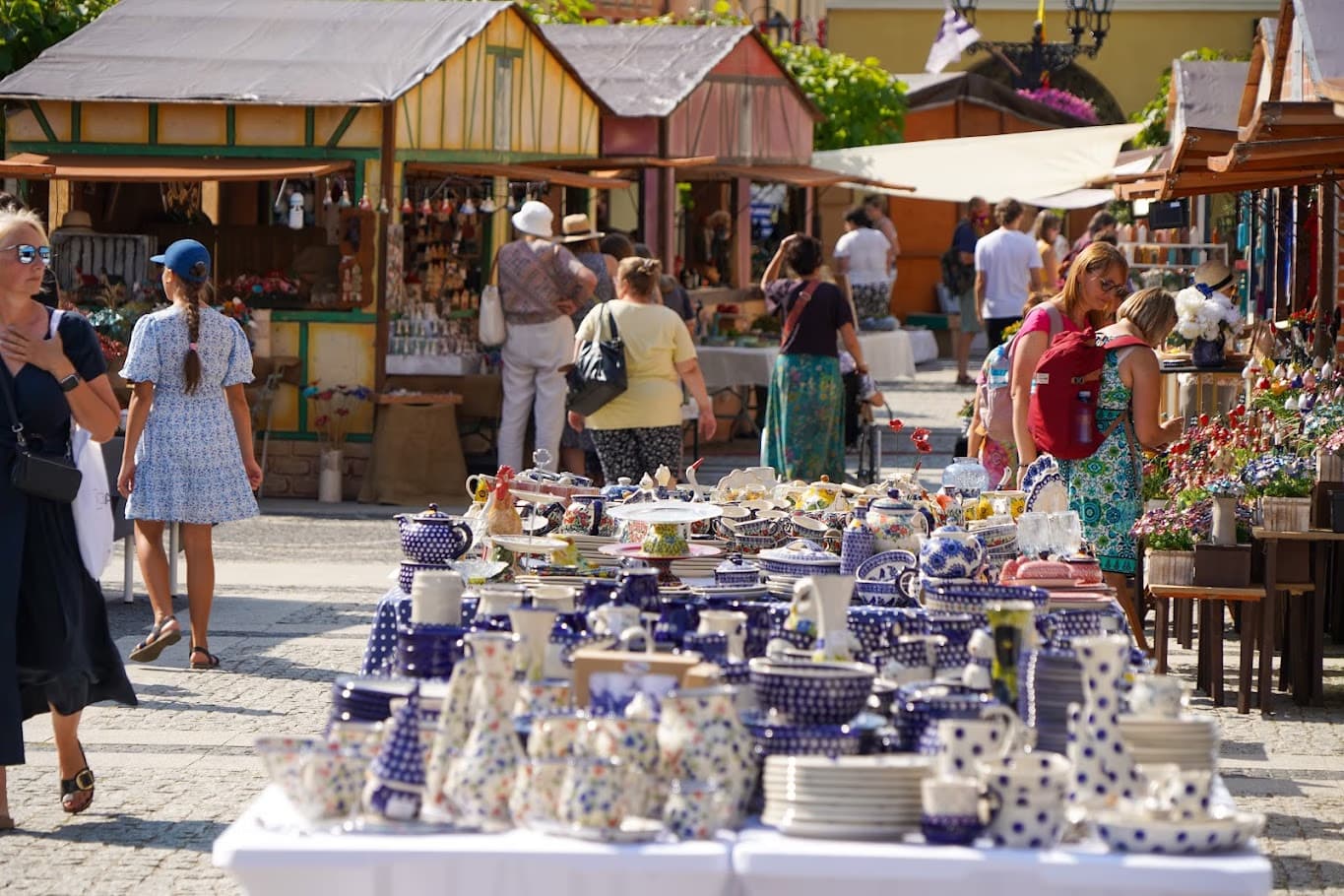 A market stall displays blue and white pottery with visitors walking among stalls in a town square.