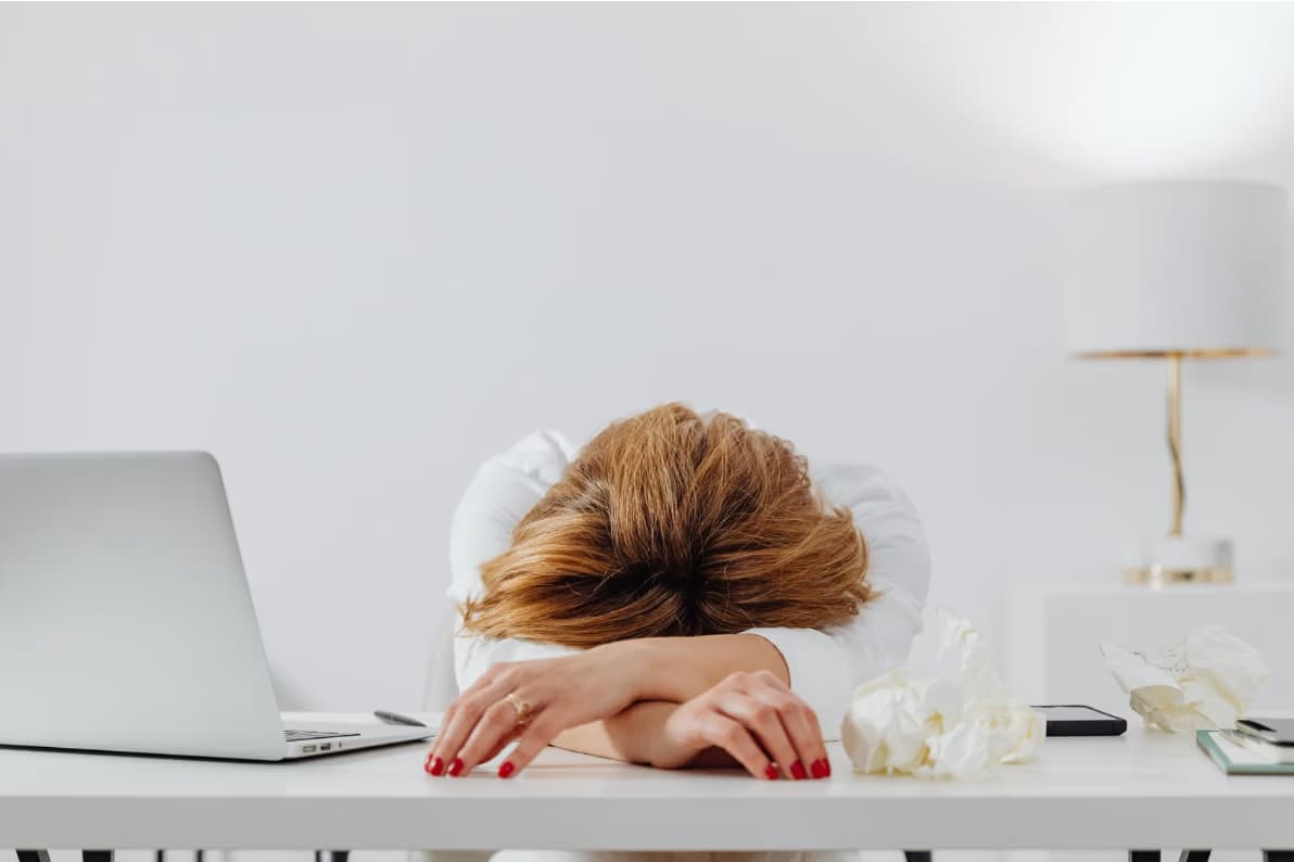 A woman rests her head on her arms at a white desk with a laptop.