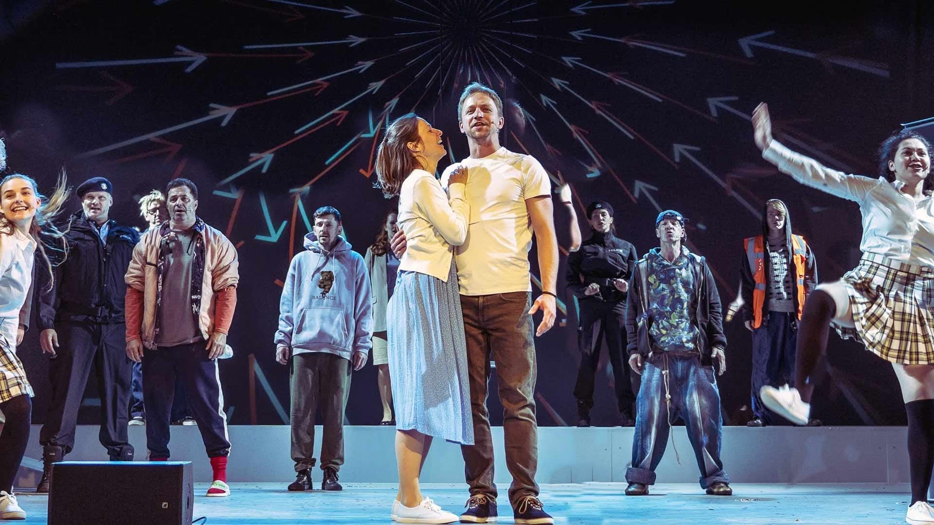 Actors performing on stage in front of a dark backdrop with radiating arrow graphics.