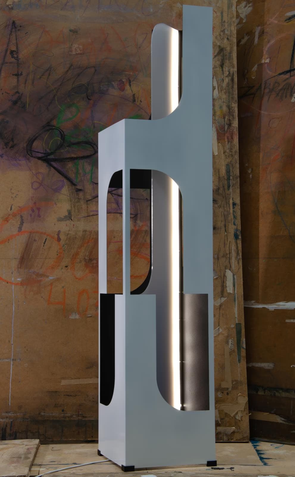 Tall, white, modern sculpture with arched cutouts stands before a graffiti-covered wooden wall.