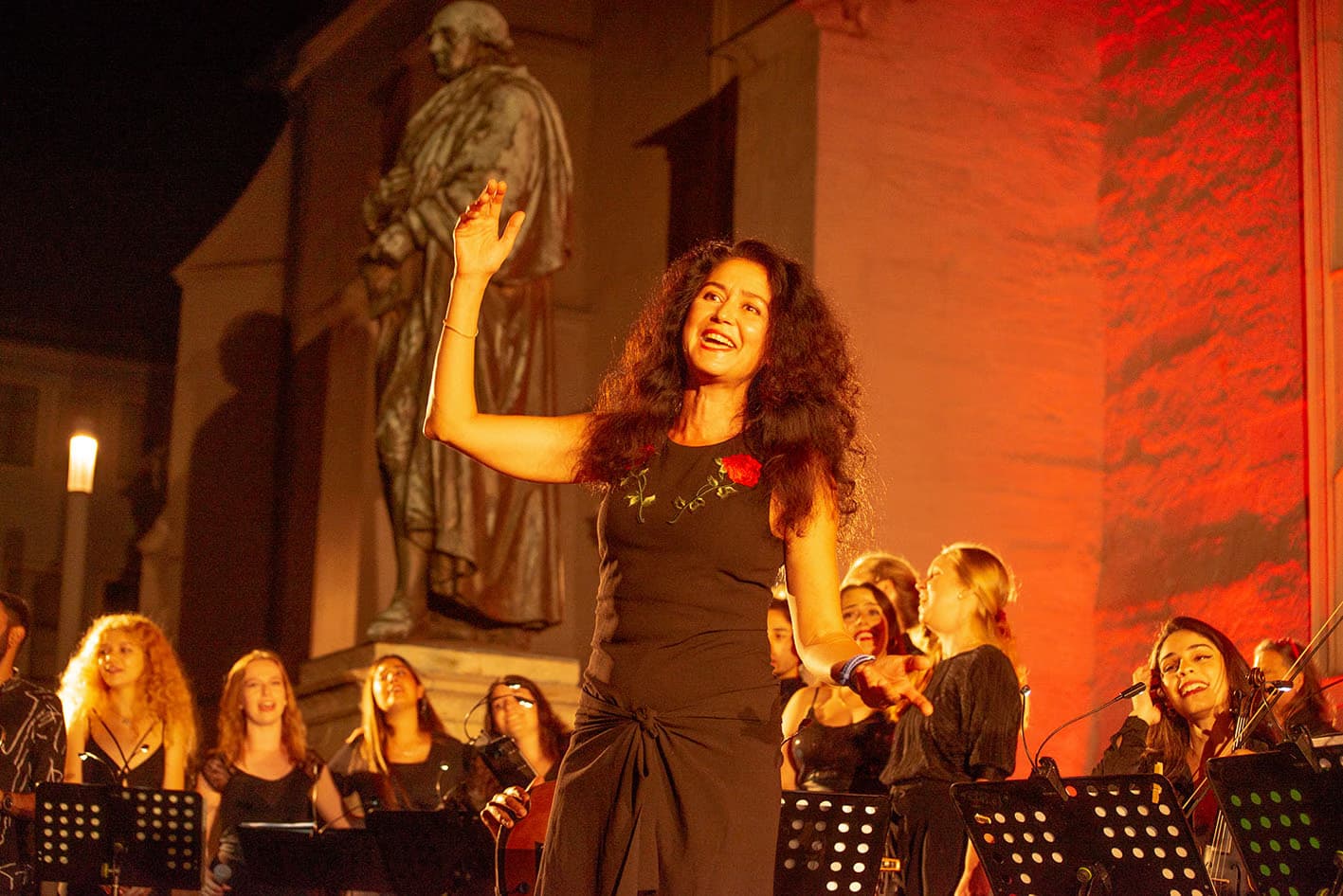A curly-haired woman sings on an outdoor stage with a choir behind her.