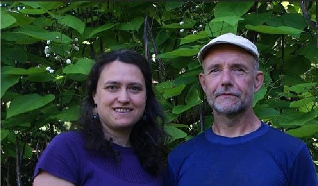 A smiling woman and a man in a cap stand before lush green foliage.