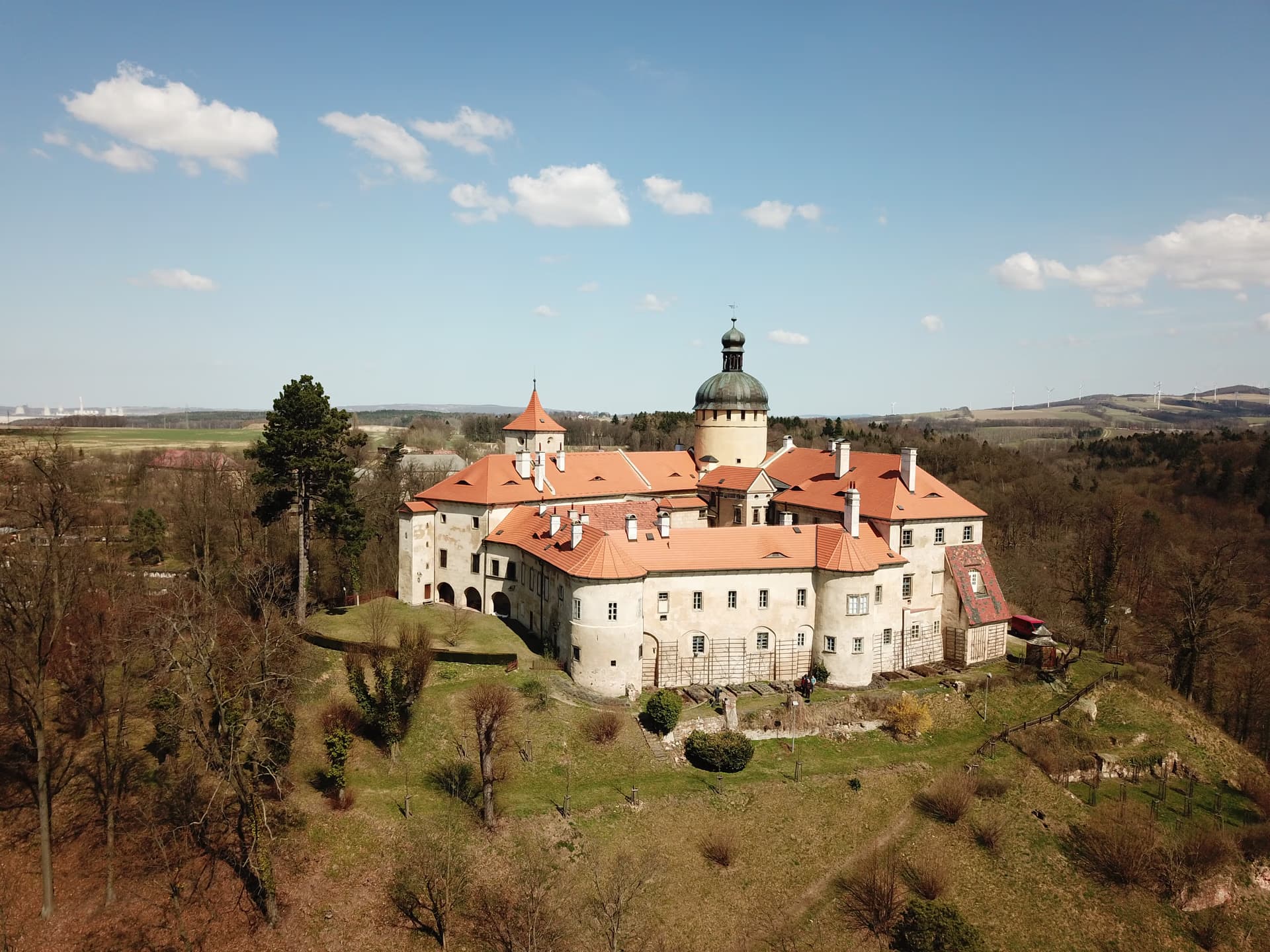 Aerial view of a historic castle with red-tiled roofs nestled on a hillside among bare trees.