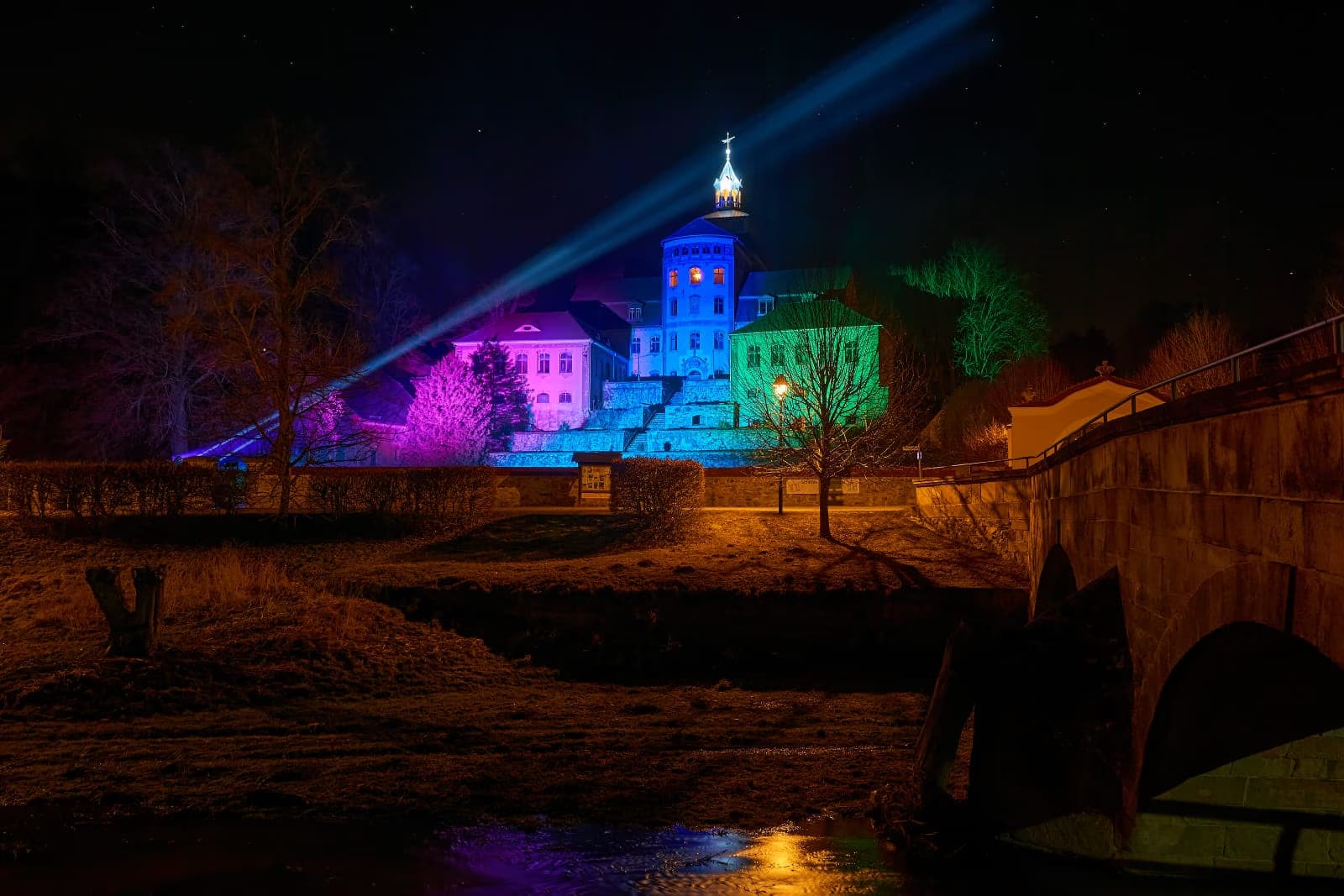 A historic castle illuminated by colourful night lights with a bright spotlight beam above.