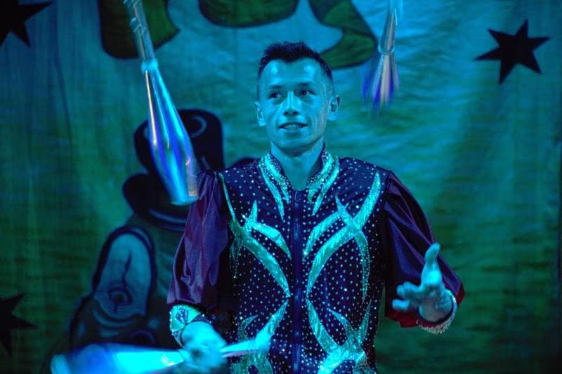 A circus juggler in a sparkling costume performs with clubs against a blue backdrop with star designs.