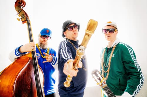 Three men in tracksuits and sunglasses pose with a double bass, a microphone, and a guitar.