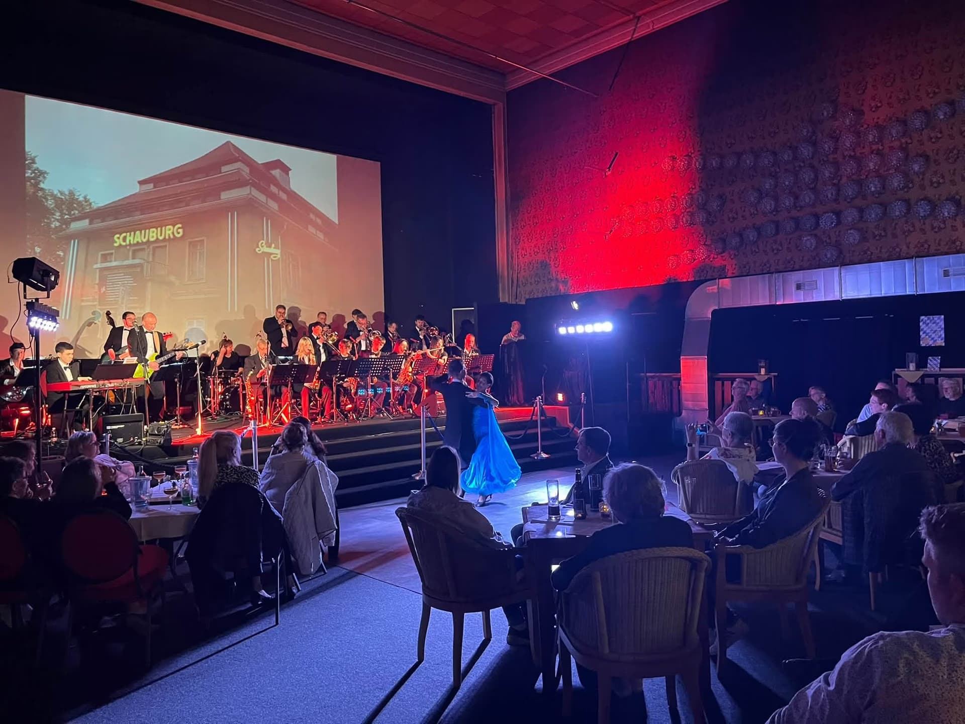 A live orchestra performs on stage before an audience in a dimly lit concert hall.
