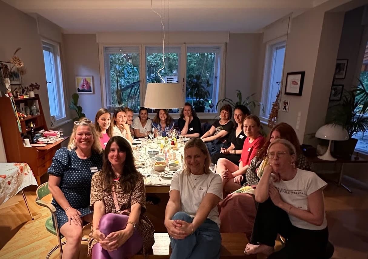 A group of women are smiling around a cozy dining table in a warm, lit room.