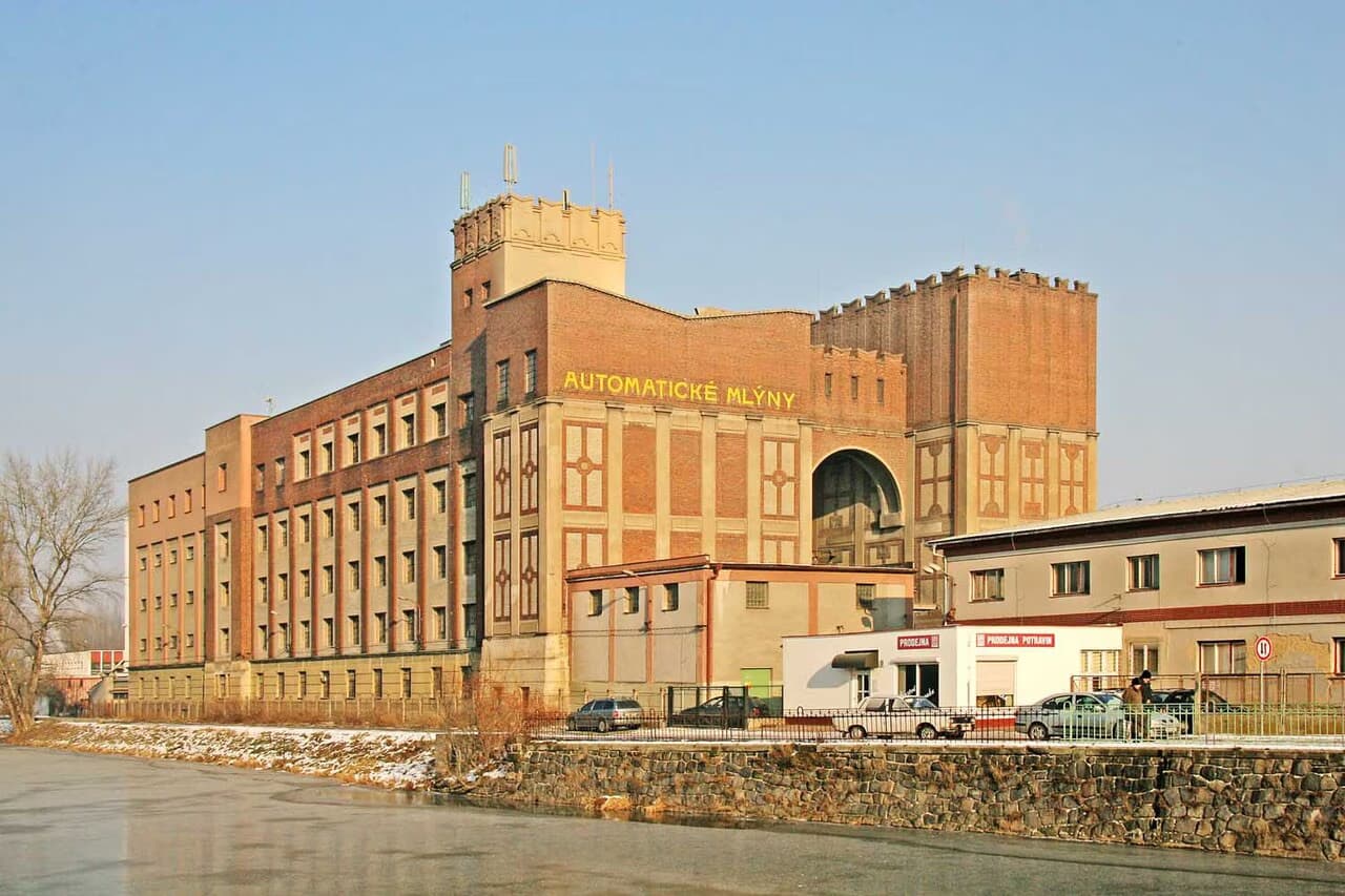 The historic Automatické Mlýny mill building stands prominently along a riverside bank under a clear sky.