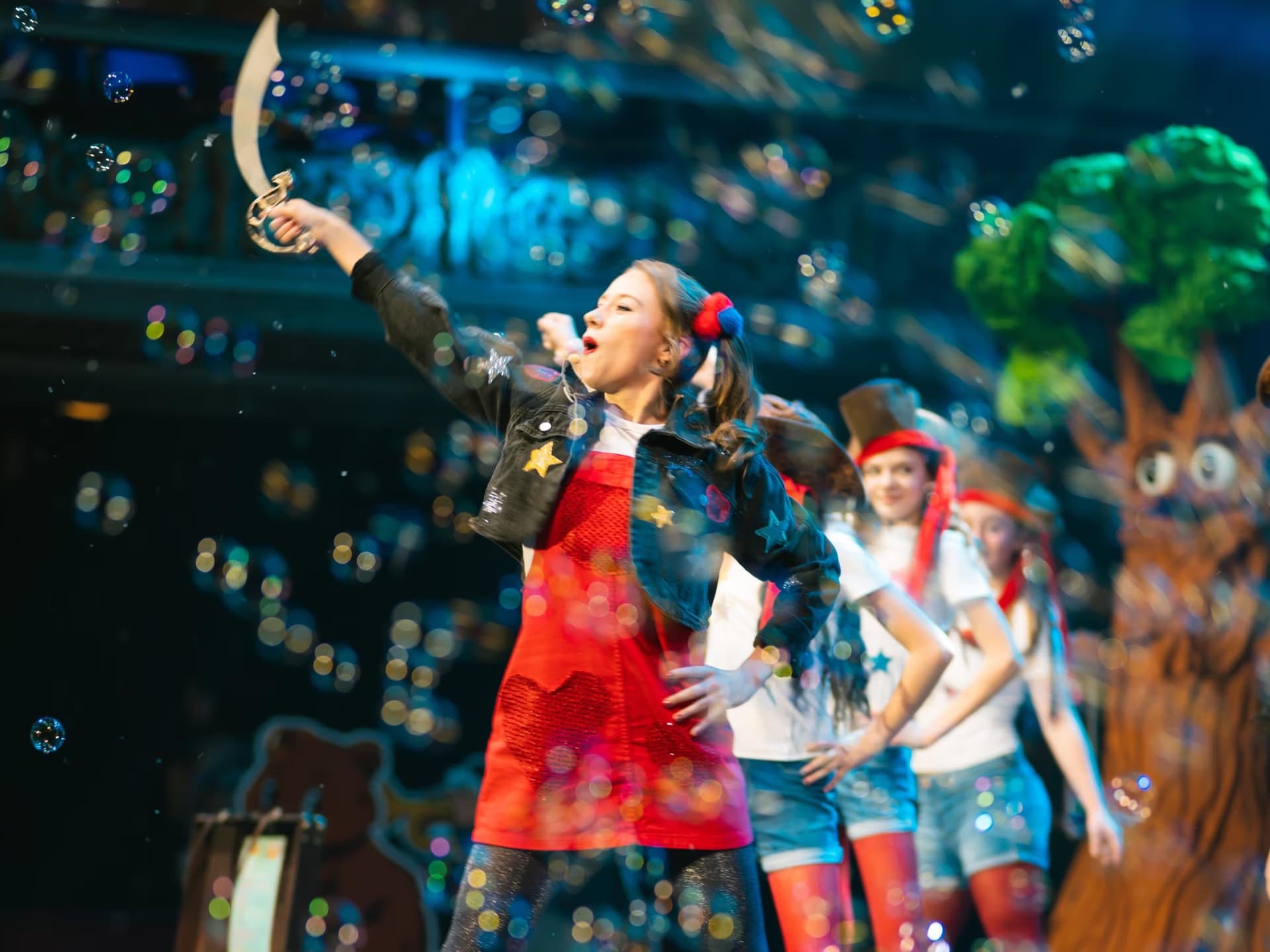 A young woman on stage holds a toy sword while bubbles float around performers and a tree prop.
