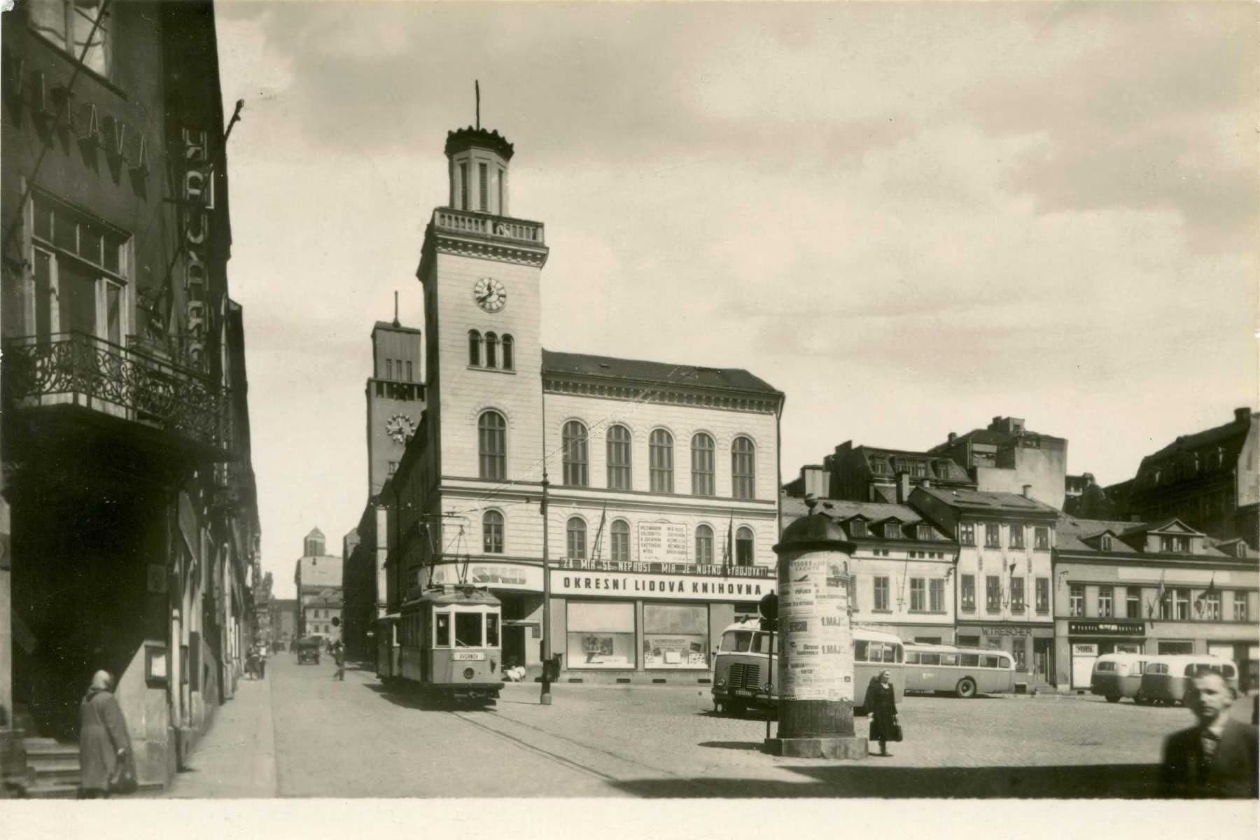 A vintage street scene featuring a town hall building with a tower, a tram, and people in an old square.