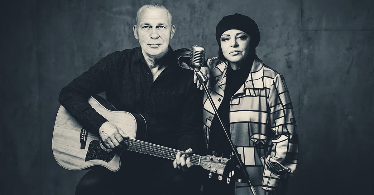 A bald man with an acoustic guitar standing next to a woman in a beret by a microphone.