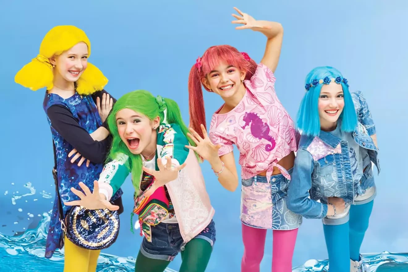 Four girls wearing brightly colored wigs smile and pose against a blue aquatic-themed background.
