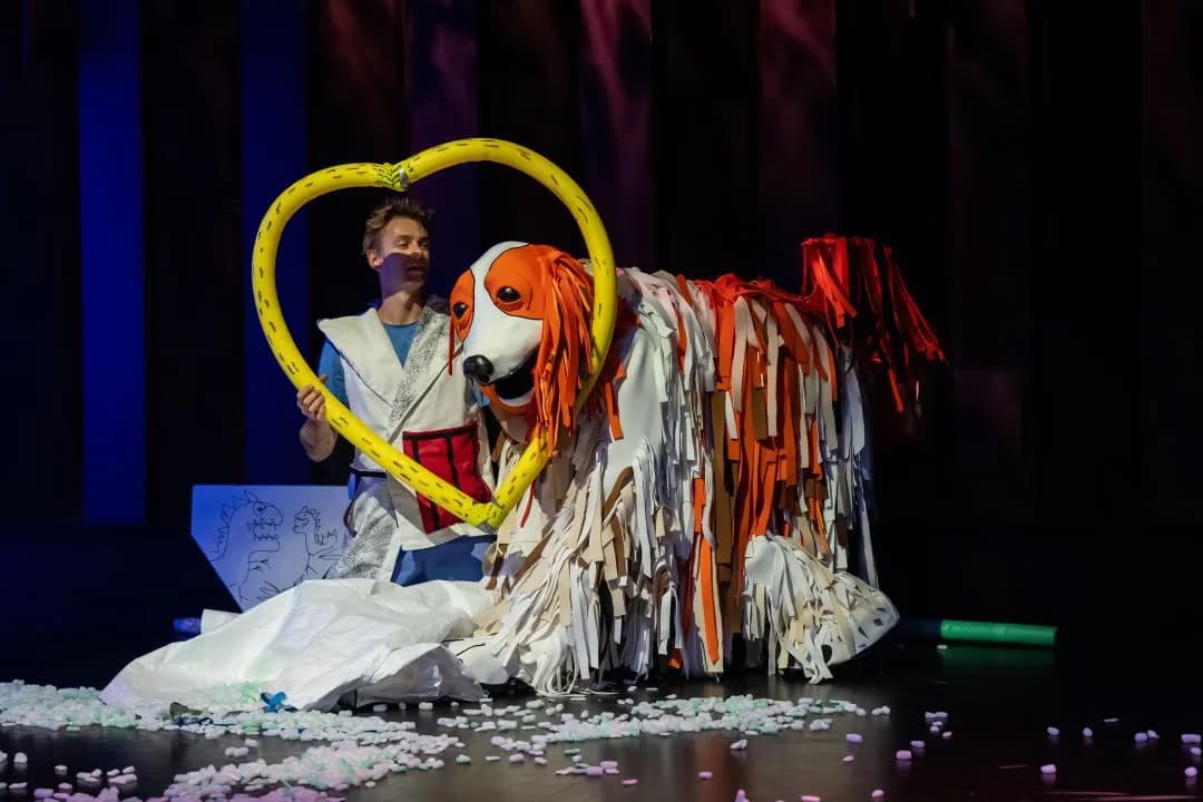 A performer holds a yellow heart frame around a large, shaggy dog puppet on a stage.