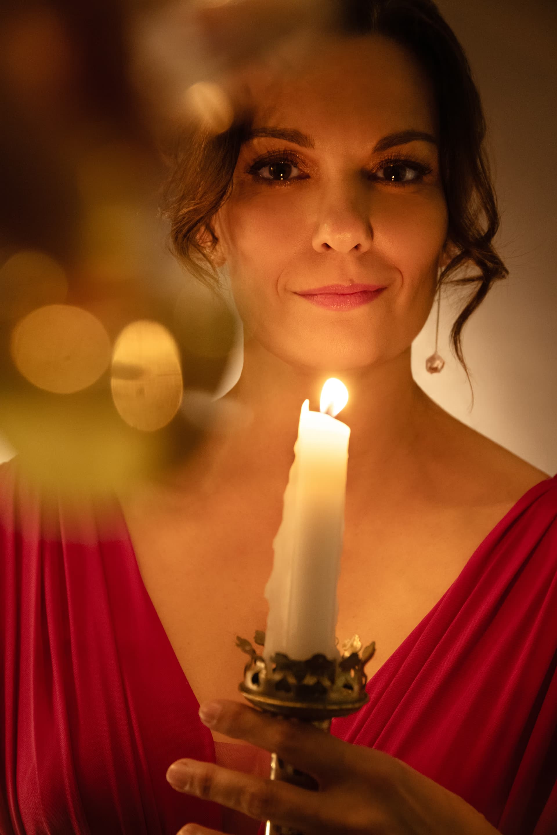 A woman in a red dress holds a lit candle, illuminated by warm, soft light.