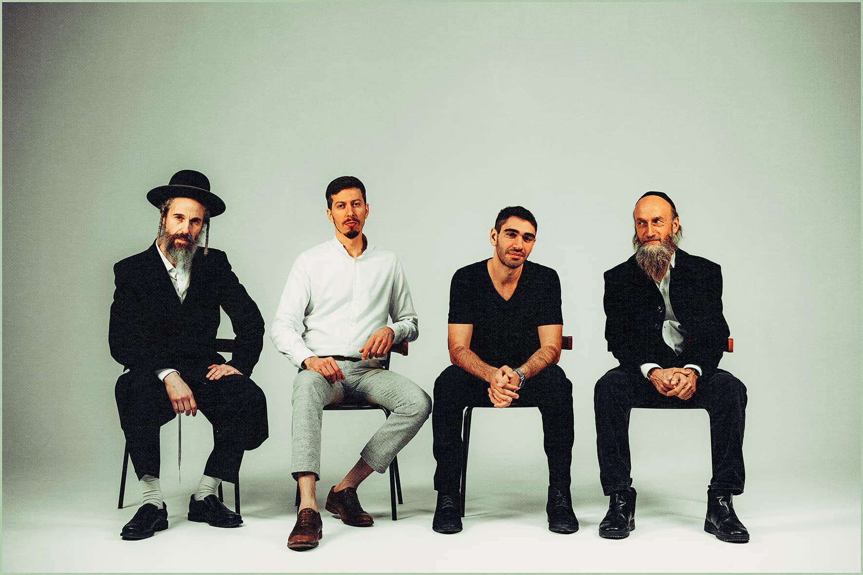 Four men sitting on simple chairs against a plain light background.