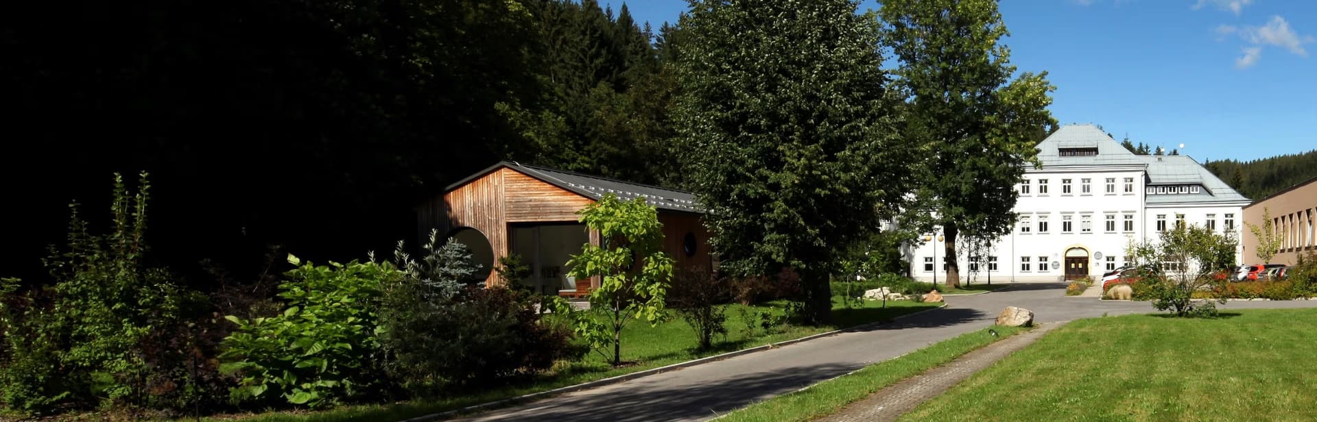 A white building stands behind green trees and a small wooden structure with a sunny lawn.