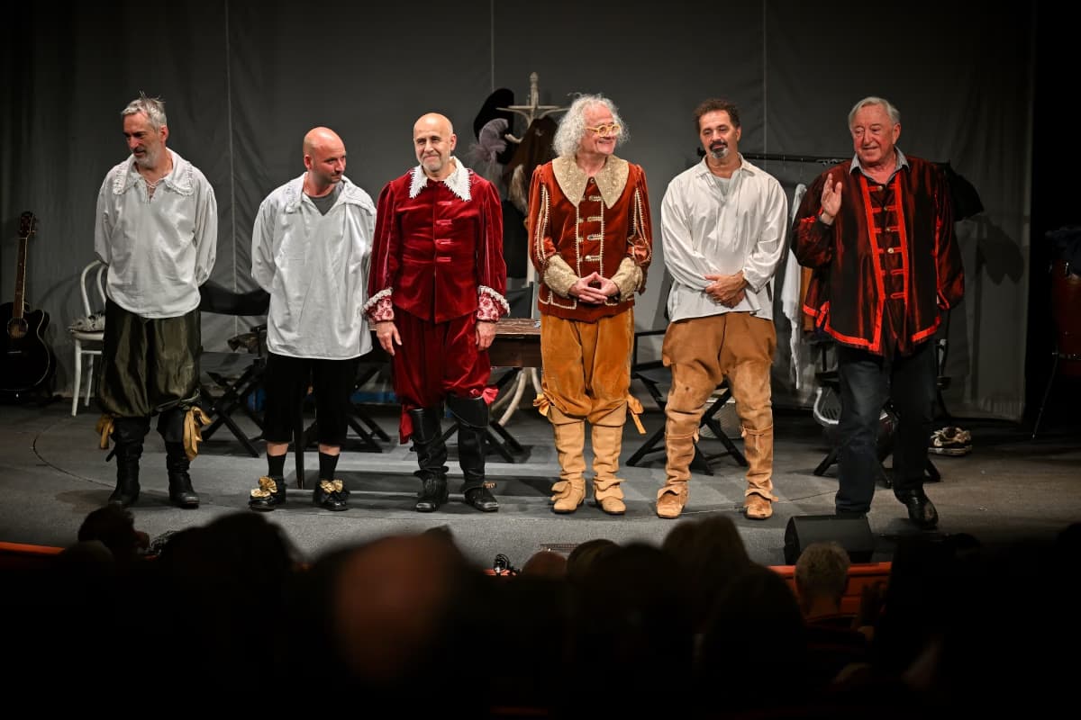 Sześciu mężczyzn w historycznych kostiumach stoi razem na scenie teatralnej przed publicznością.