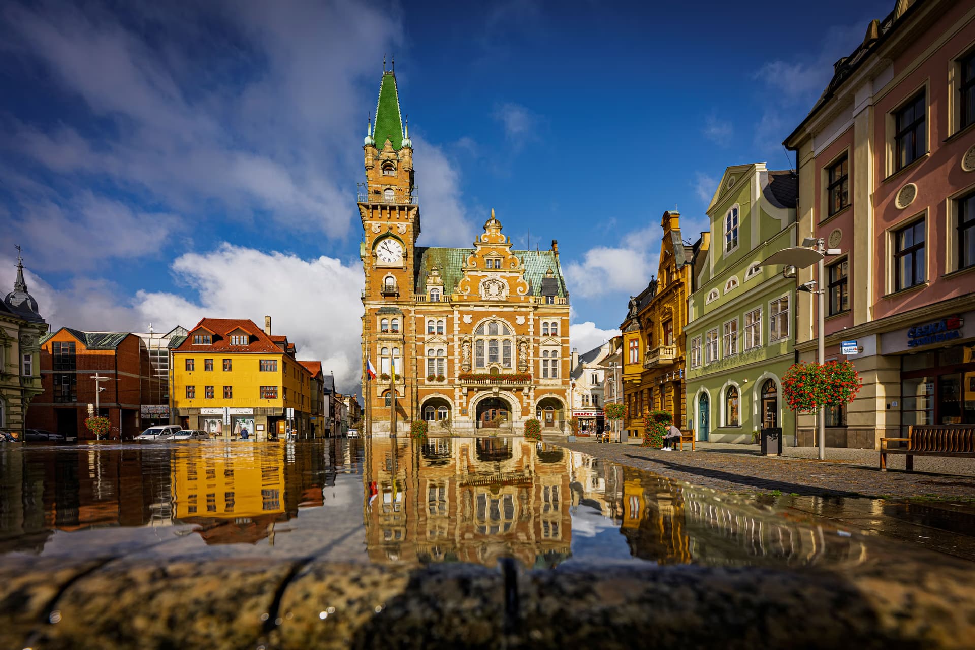 A historic town hall with a green clock tower reflects in the water of a city square.