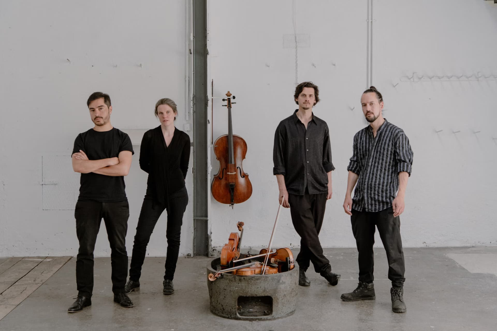 Four musicians stand in a minimalist room with a cello on the wall and instruments in a pile.