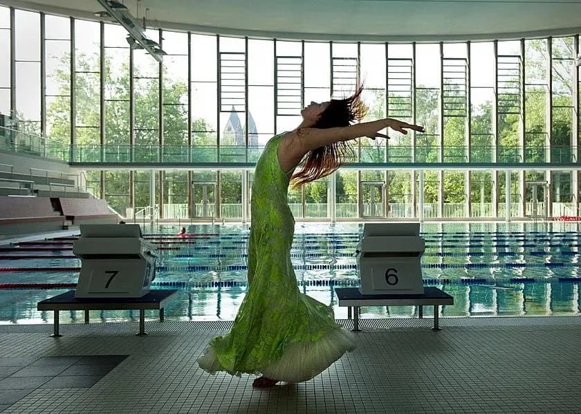 A woman in a vibrant green dress poses dramatically on the deck of an indoor swimming pool.