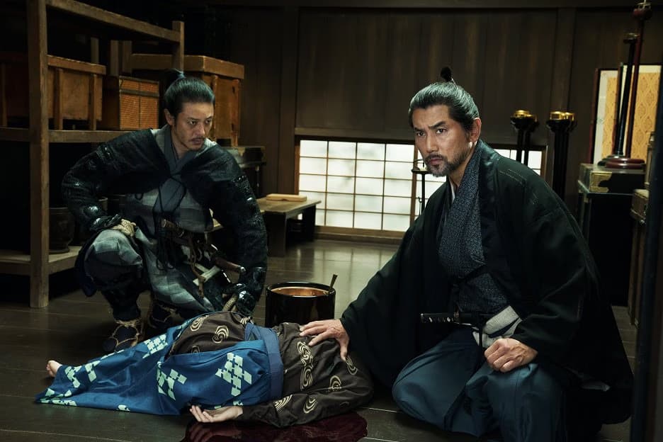 Two men in traditional Japanese clothing observe a person lying on the floor in a dimly lit room.