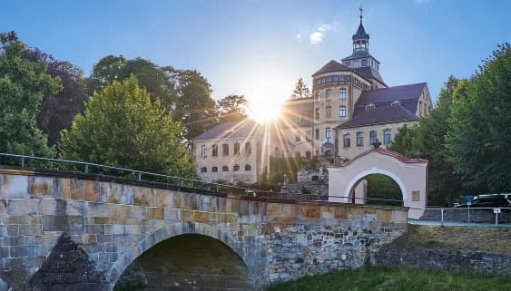 A historic stone castle stands behind a sunlit stone bridge with an arched gateway.