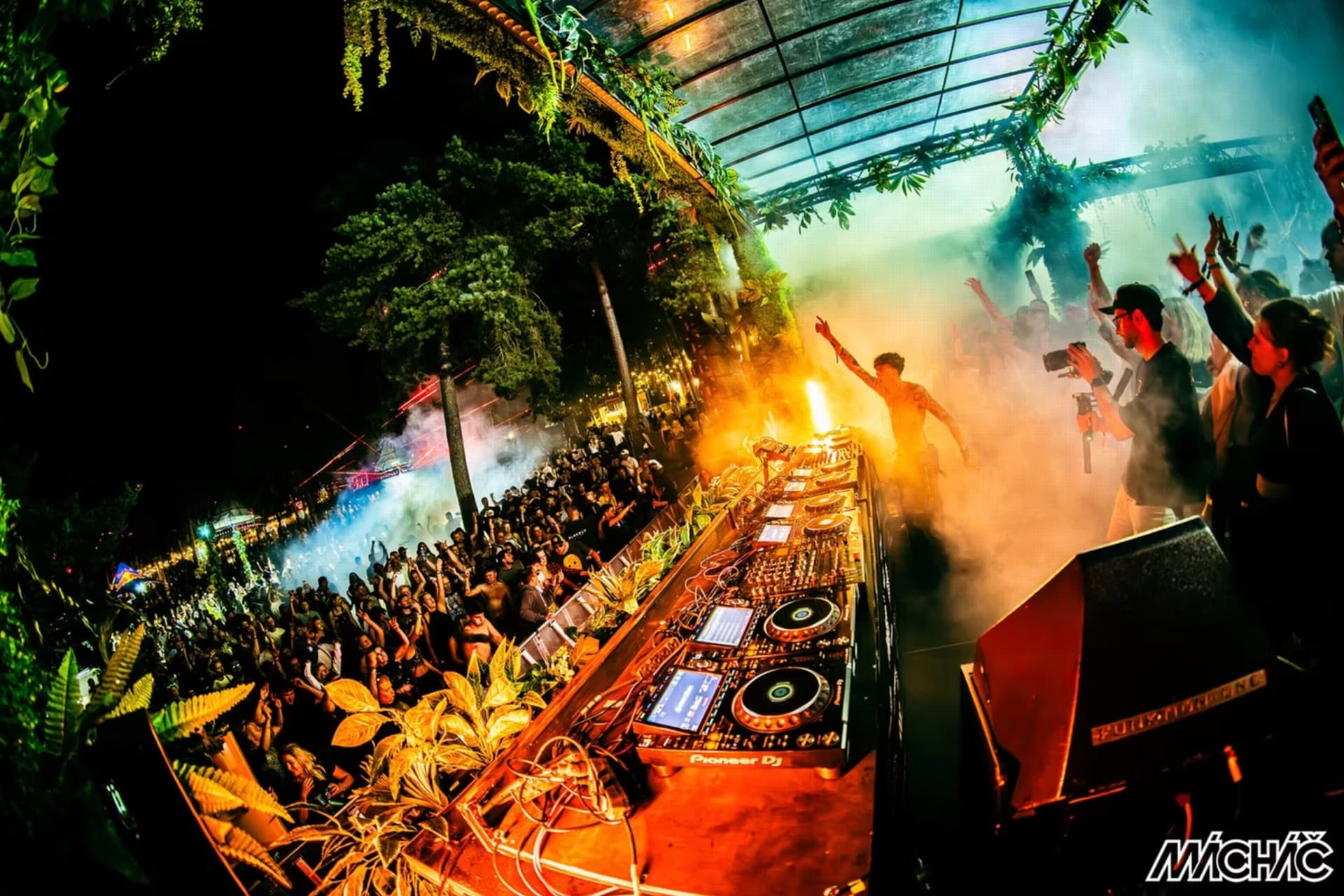 A nighttime outdoor music festival with a DJ booth, dancing crowd, and decorative jungle-like foliage.