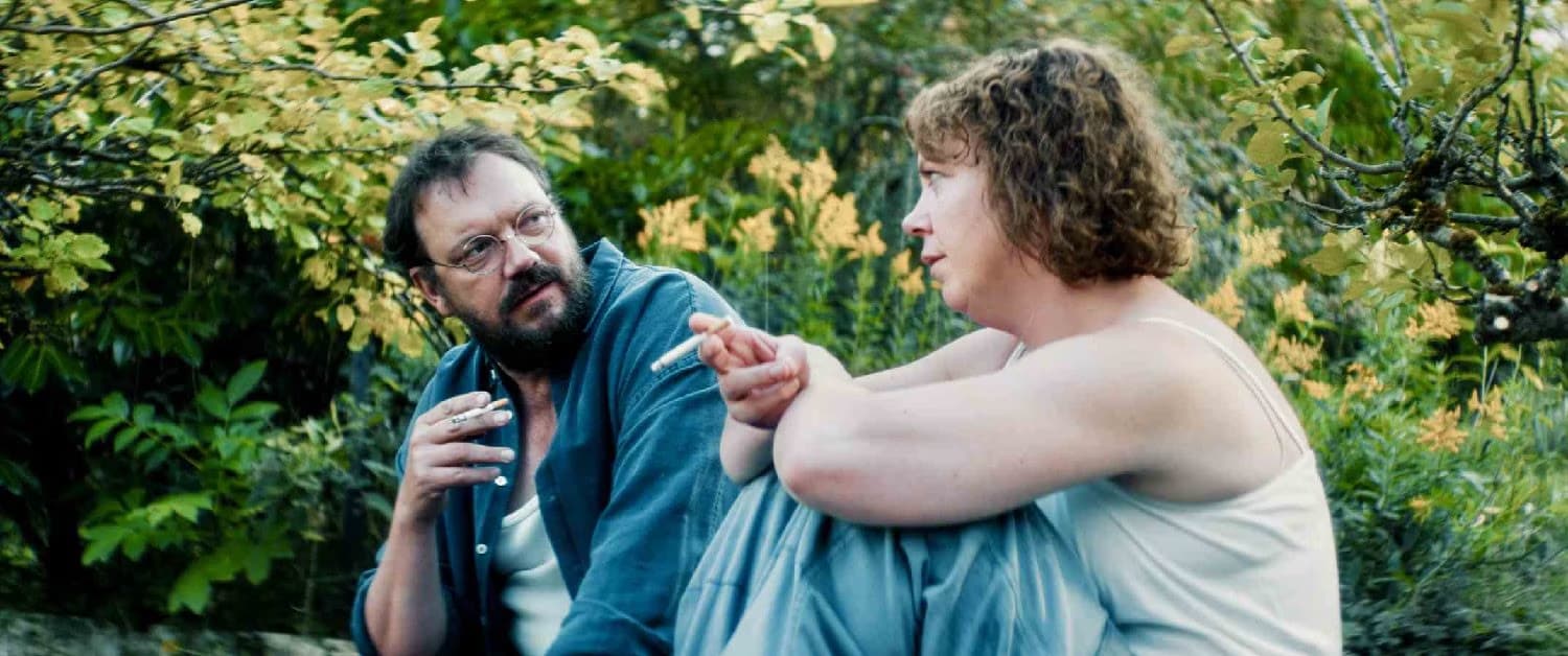A bearded man and a woman talk outdoors in a garden setting with green foliage.